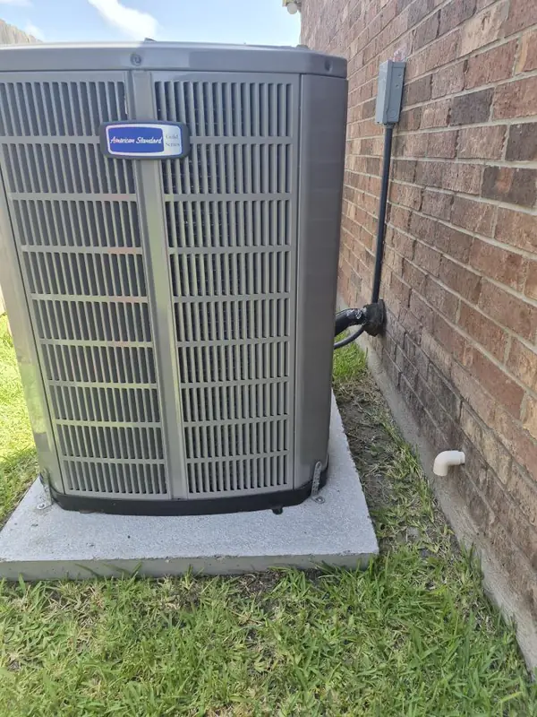 American Standard Gold outdoor unit on a concrete pad against a brick wall, with the disconnect and whip mounted beside it